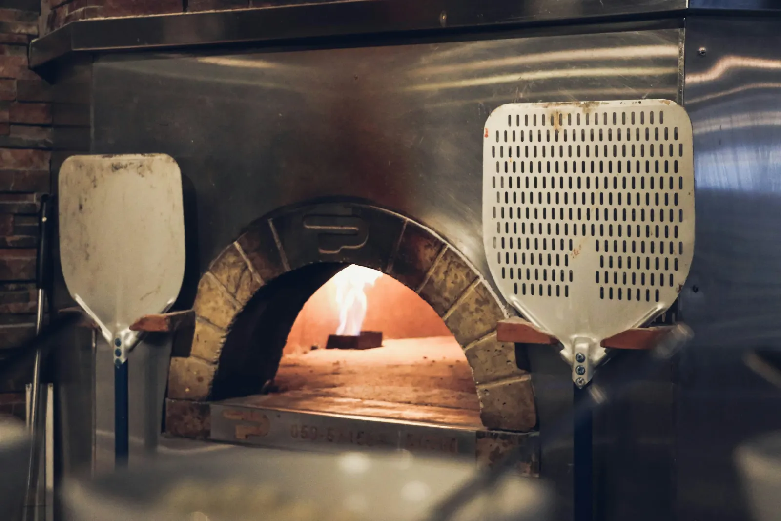 Gemauerter Holzofen mit Flamme, daneben zwei Pizzaschieber